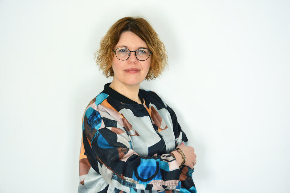 Na zdjęciu kobieta o jasnych, kręconych włosach za uszy, w kolorowej koszuli. 