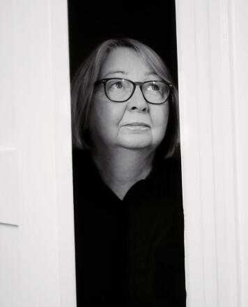 Na zdjęciu kobieta wyglądająca przez okno, ubrana w czarną koszulę, na nosie okulary.