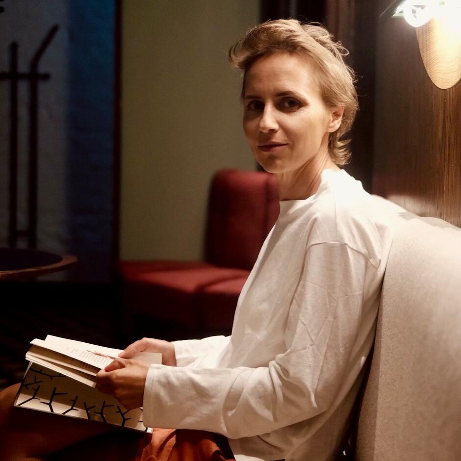 Na zdjęciu kobieta ubrana w białą koszulę, siedząca na kanapie. W rękach trzyma książkę.