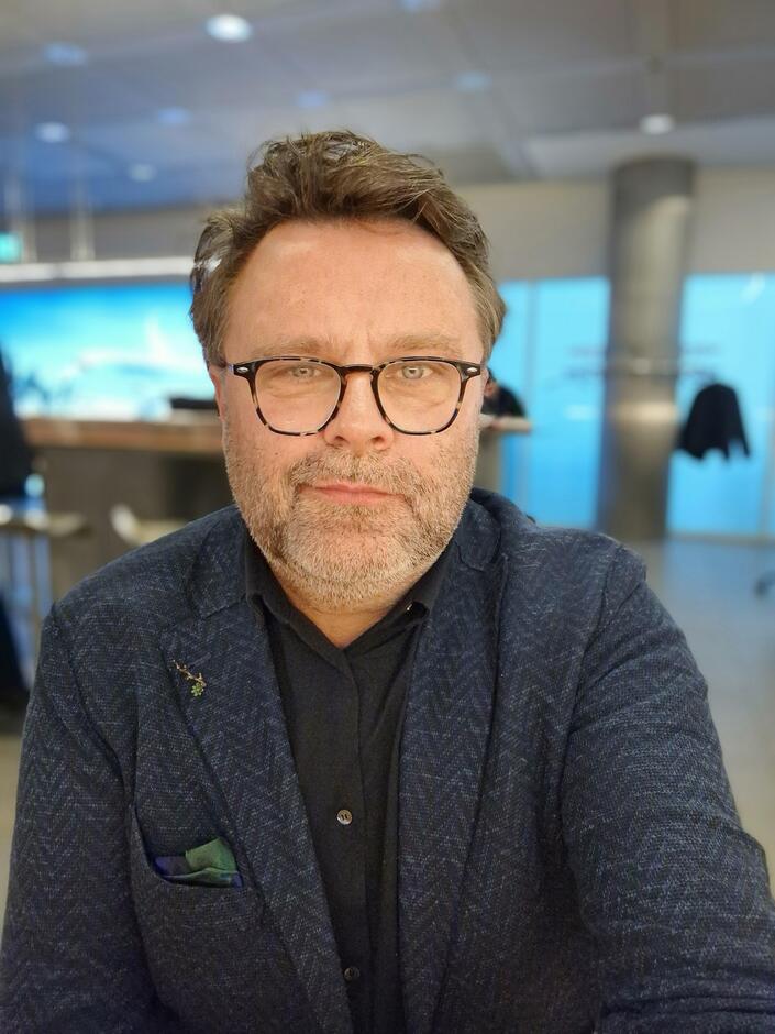 Na zdjęciu mężczyzna w okularach, z lekkim zarostem na twarzy, krótkie włosy. Ubrany w ciemną marynarkę.