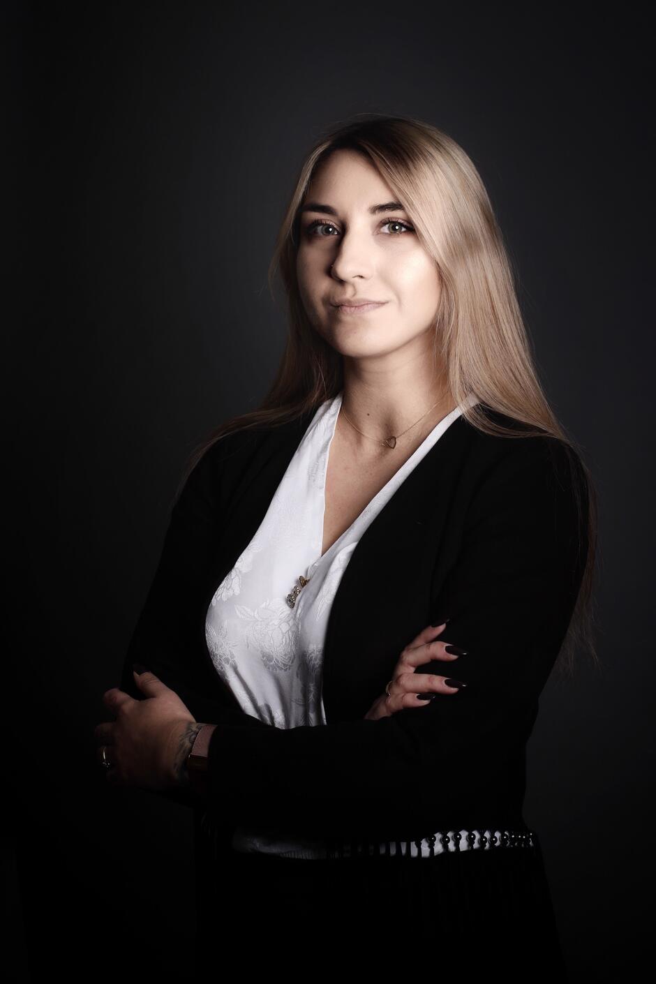 Na zdjęciu kobieta z długimi jasnymi włosami, ubrana w czarny sweter i białą koszulę.