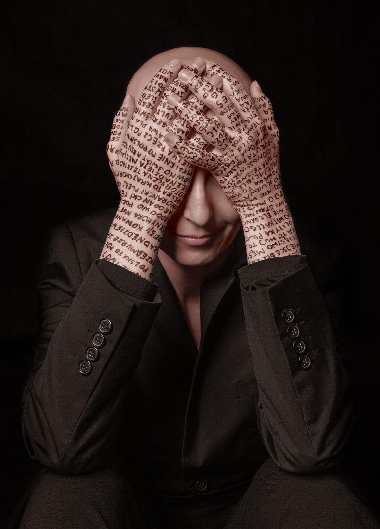 Na zdjęciu łysa kobieta, z założonymi rękoma na głowie, zakrywającymi twarz. Ręce pokryte są tatuażami.