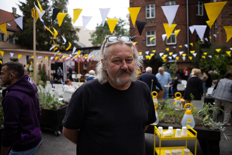 Mężczyzna z długimi siwymi włosami i wąsami pozuje do zdjęcia na dziedzińcu dawnej szkoły, za nim uczestnicy wydarzenia.