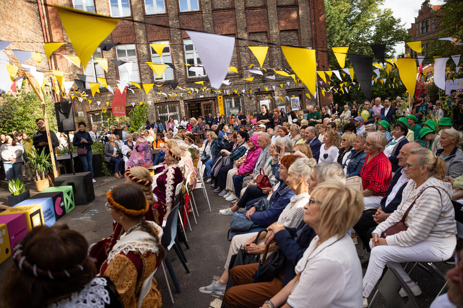 Tłum ludzi siedzi na krzesłach na dziedzińcu dawnej szkoły podczas wydarzenia.