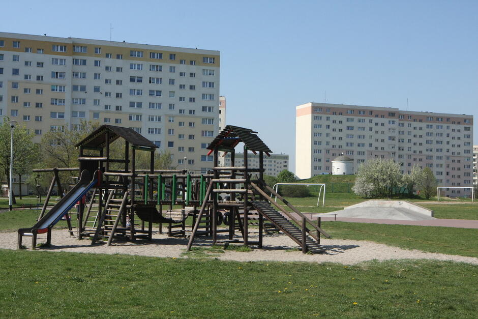 na pierwszym planie drewniana konstrukcja dla dzieci na placu zabaw, w tle dwa wysokie bloki mieszkalne