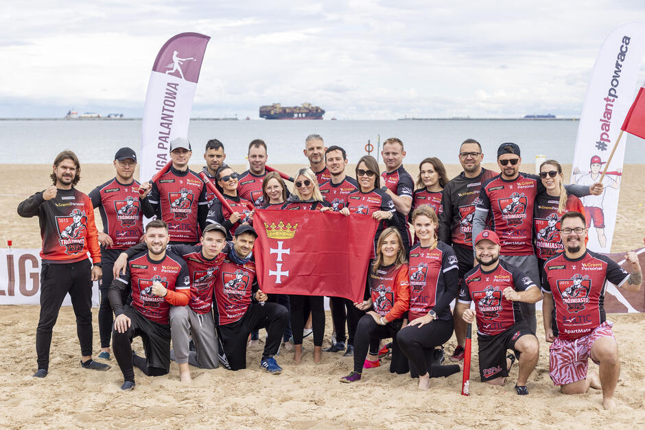 Grupa ponad 20 osób w strojach sportowych. Pozują do wspólnego zdjęcia, centralnie trzymają flagę Gdańska