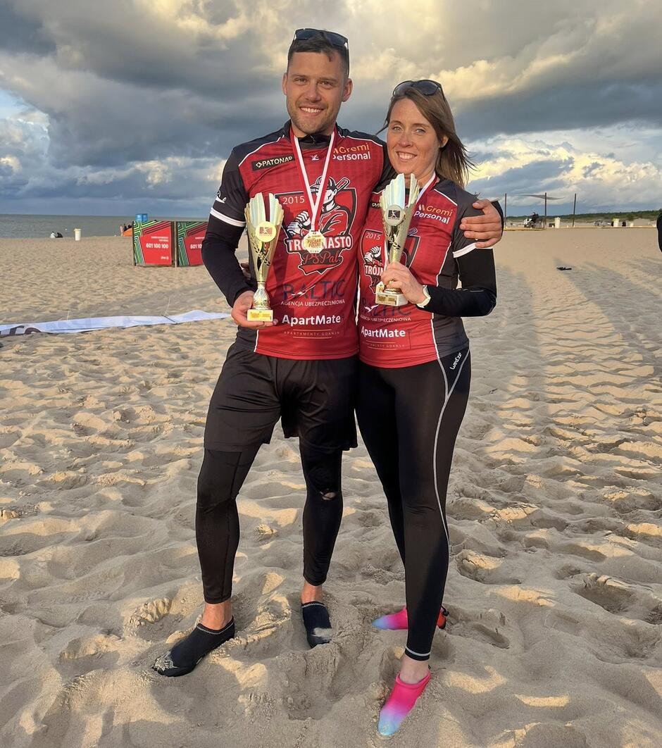 Dwie osoby w strojach sportowych, pozują do zdjęcia, trzymając w ręku puchary zwycięzców. Po lewej mężczyzna, po prawej - kobieta