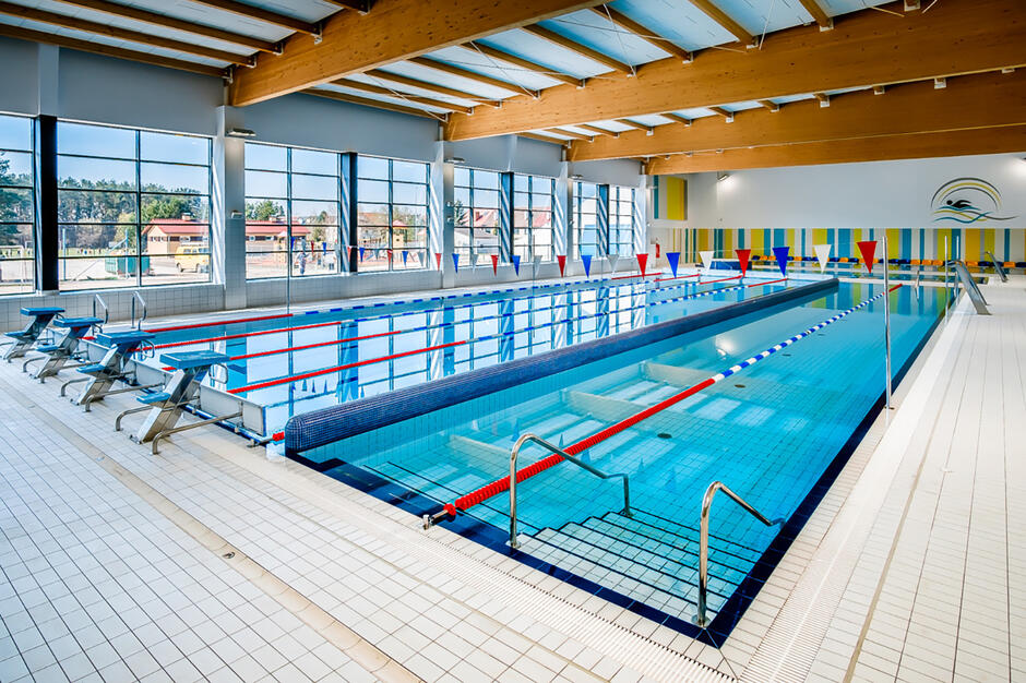 Pusta pływalnia z kilkoma torami do pływania, drewnianym dachem i dużym oknem, przez które widać zabudowę osiedla mieszkaniowego.