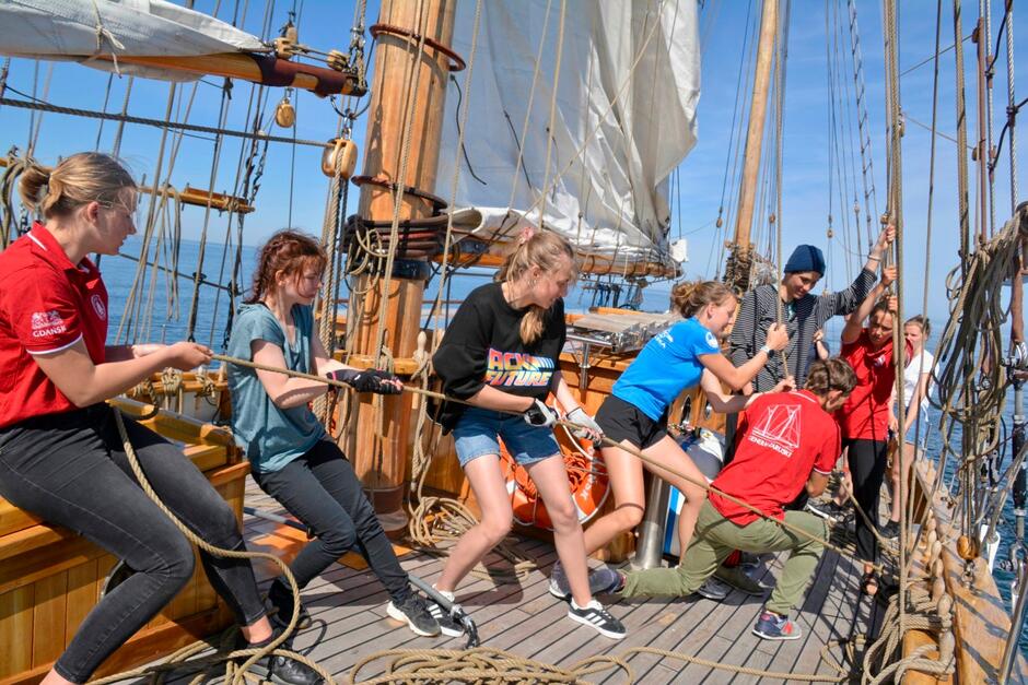 Młodzież pracuje na pokładzie żaglowca. Kilka osób ciągnie linę.