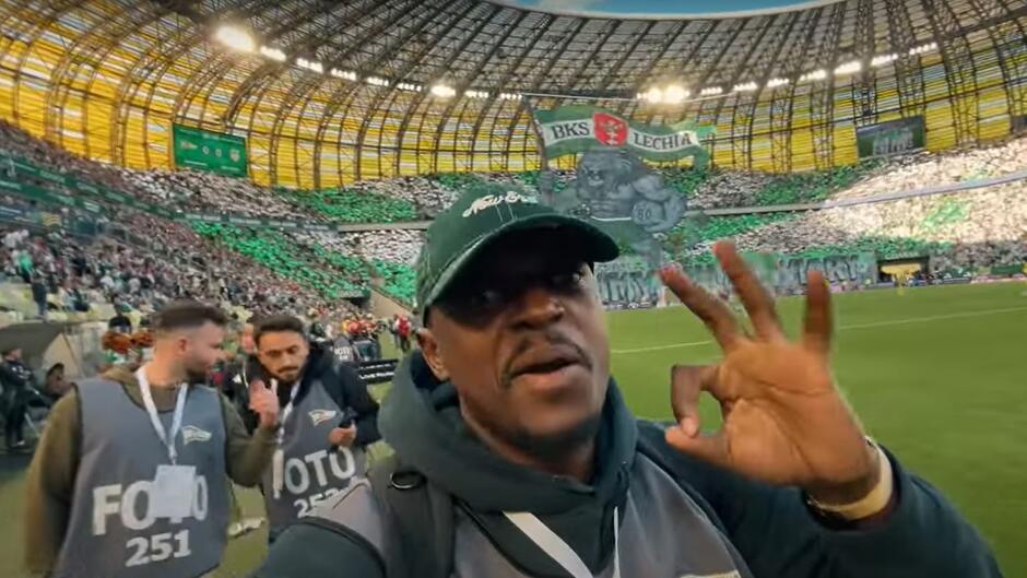 Mężczyzna na stadionie podczas meczu, wykonuje dłonią gest typu Prima sort. Za jego plecami widać trybuny stadionu, zapełnione kibicami