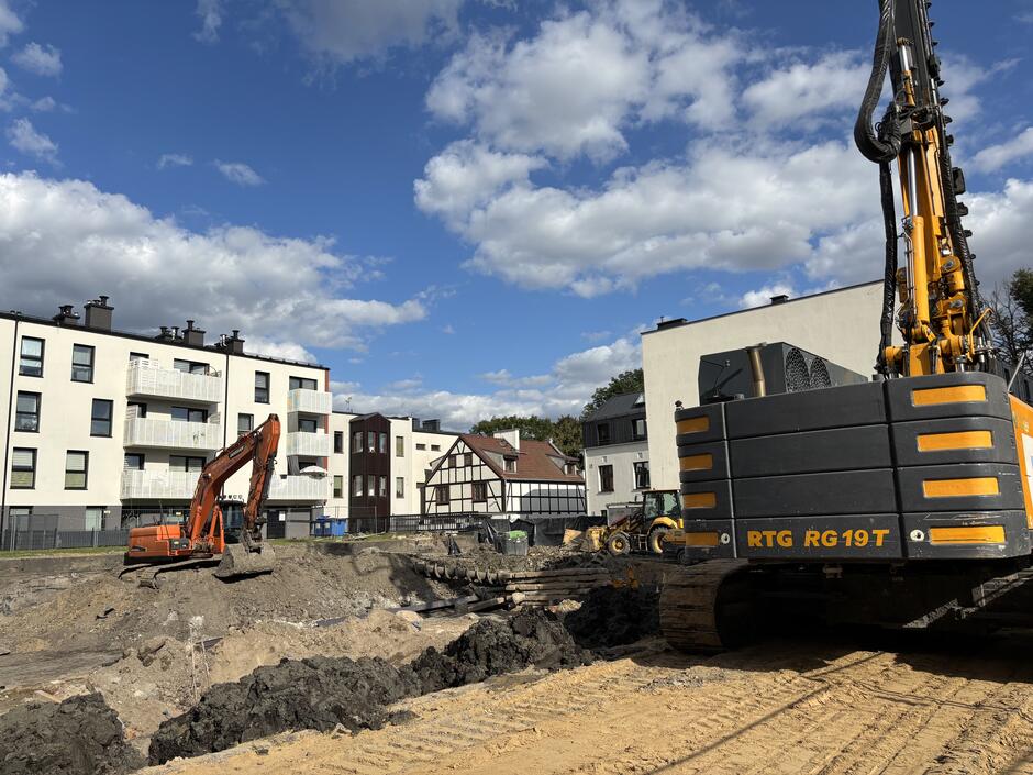 Dwie koparki na rozkopanej ziemi między kamienicami