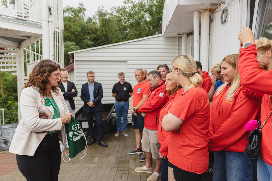 Kobieta w garsonce stoi przed grupa ubranych na czerwono ratowników wodnych i wręcza im upominki.