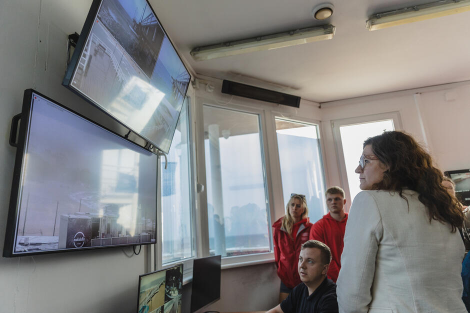 Mężczyzna na krześle siedzi przed monitorami, na których widać plażę. Obok stoi kobieta i obserwuje monitory.