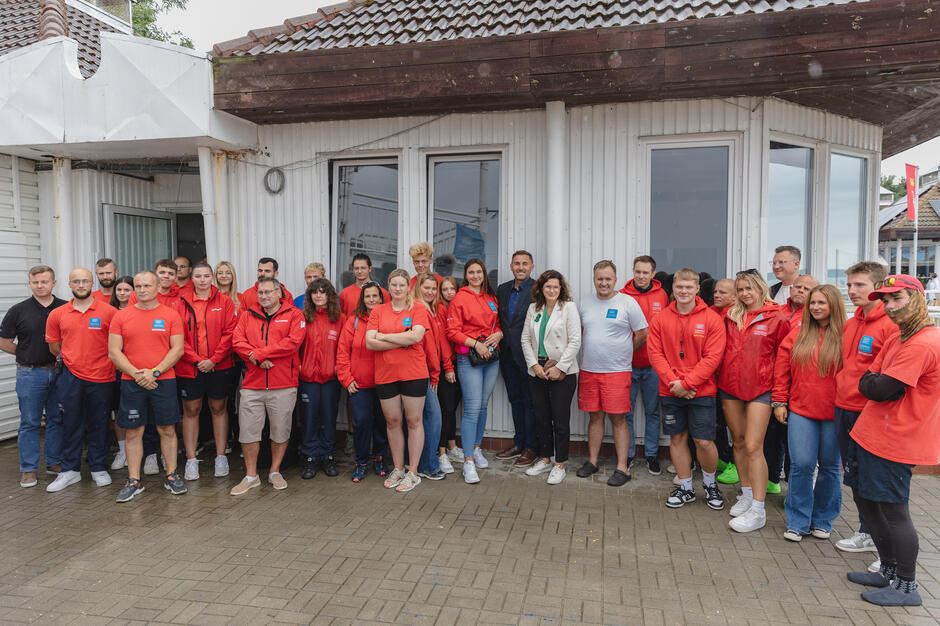 Liczna grupa ratowników wodnych w czerwonych uniformach pozuje do zdjęcia pod obiektem na plaży z władzami miasta.
