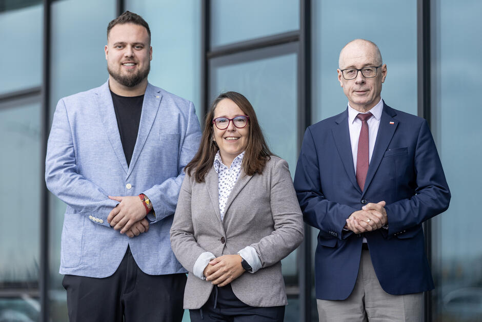 Trzy osoby stoją, pozując do wspólnego zdjęcia. Kolejno: mężczyzna, kobieta, mężczyzna 