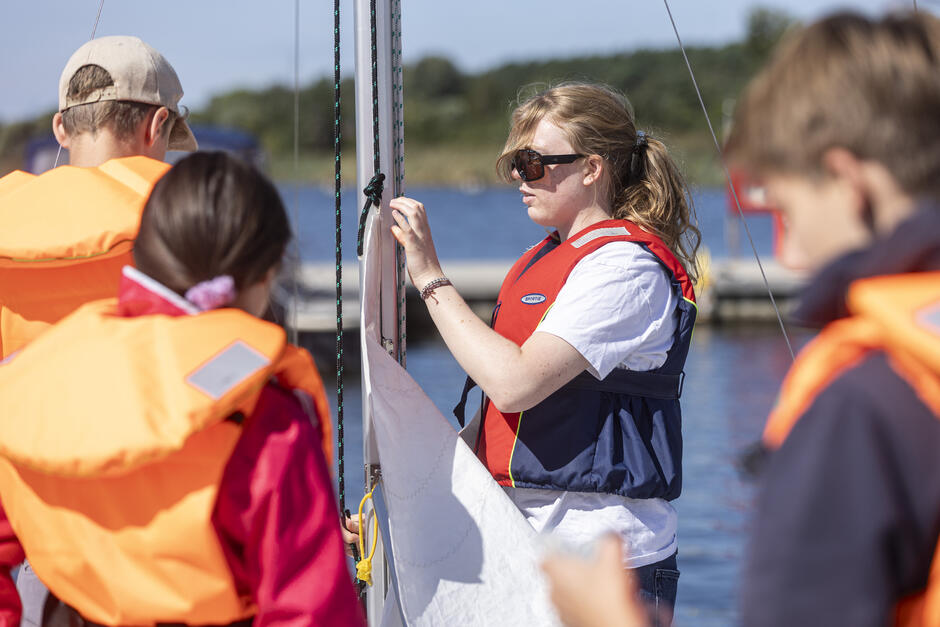 na zdjęciu młoda dziewczyna przyczepia żagiel do masztu, obok niej trzy osoby w pomarańczowych kapokach