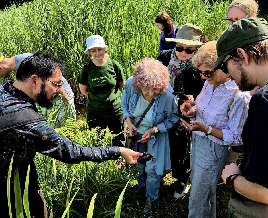 Grupa osób ogląda coś, co pokazuje im w ręku mężczyzna. Są otoczeni trawami podczas spaceru przyrodniczego.