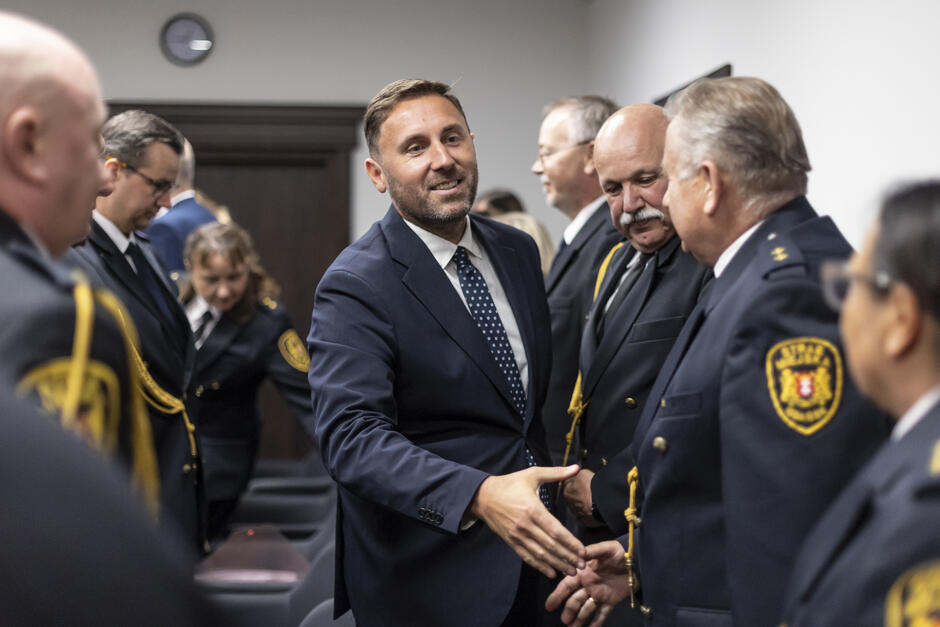 Mężczyzna w garniturze ściska rękę, śmiejąc się strażnikowi miejskiemu w sali, gdzie zebrało się wielu strażników miejskich.