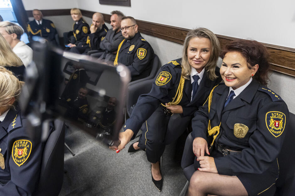 Dwie strażniczki miejskie śmieją się, robiąc sobie zdjęcie telefonem w sali, gdzie siedzi więcej strażników miejskich.