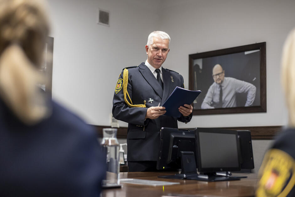Komendant Straży Miejskiej stoi z teczką w rękach w sali urzędu. Przed nim siedzą strażniczki miejskie widziane rozmyte od tyłu.