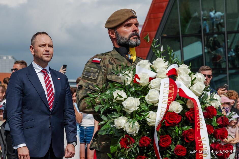 Stoją dwaj mężczyźni - jedni za drugim, zwróceni w prawo. Pierwszy - w mundurze - trzyma na rękach wieniec kwiatów z biało-czerwoną kokardą. Drugi - w garniturze - podąża za mężczyzną z kwiatami