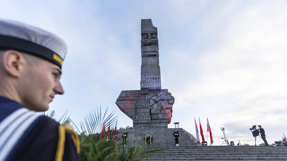 Na zdjęciu widać Pomnik Obrońców Wybrzeża na Westerplatte, przy którym stoją wartownicy i ustawiono biało-czerwone flagi. Na pierwszym planie znajduje się sylwetka marynarza w mundurze, patrzącego w stronę monumentu.
