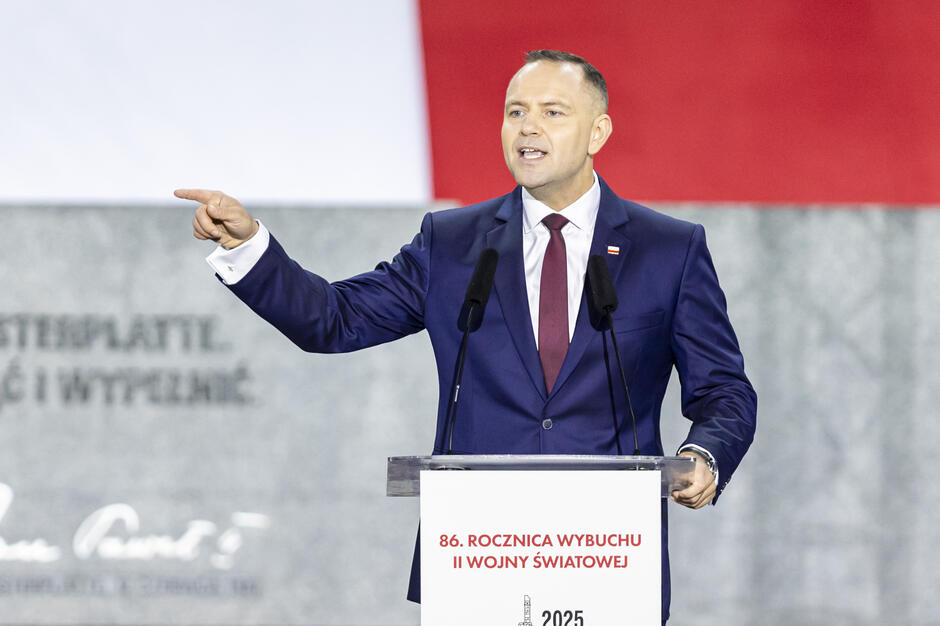 Na zdjęciu widać mężczyznę w garniturze i bordowym krawacie, przemawiającego zza mównicy z mikrofonami, z uniesioną ręką w geście podkreślającym słowa. Na mównicy widnieje napis informujący o 86. rocznicy wybuchu II wojny światowej.
