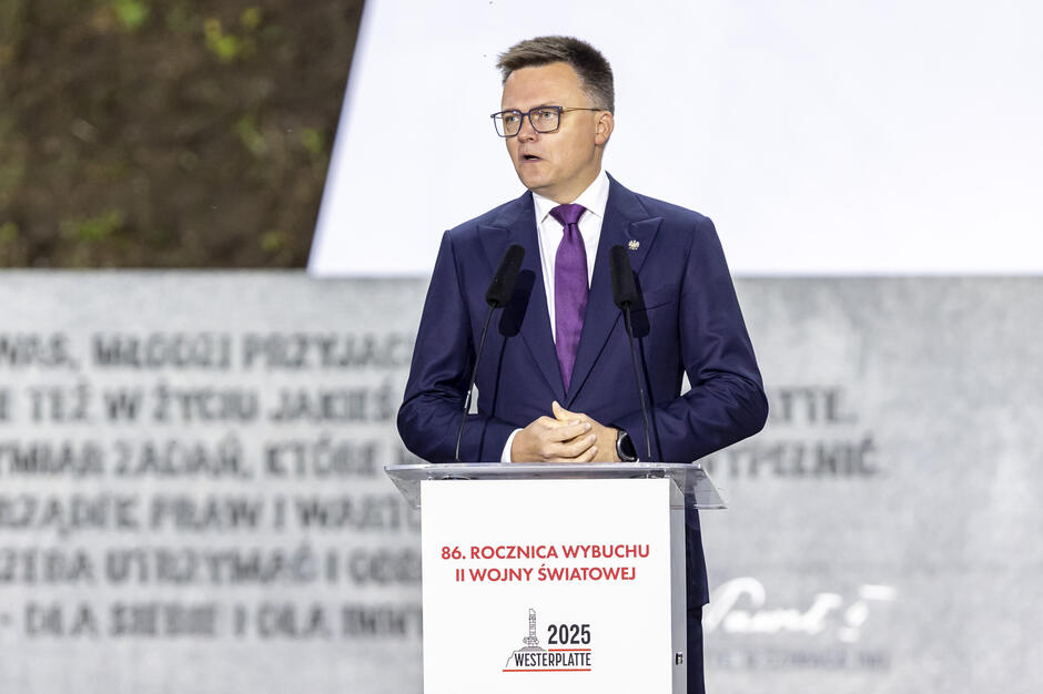 Na zdjęciu widać mężczyznę w garniturze i bordowym krawacie, przemawiającego zza mównicy z mikrofonami, z uniesioną ręką w geście podkreślającym słowa. Na mównicy widnieje napis informujący o 86. rocznicy wybuchu II wojny światowej.
