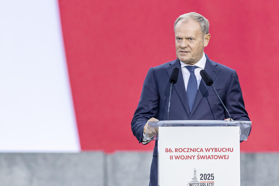 Na zdjęciu widać mężczyznę w garniturze i niebieskim krawacie przemawiającego zza mównicy z mikrofonami, na tle biało-czerwonej flagi. Na mównicy znajduje się napis informujący o 86. rocznicy wybuchu II wojny światowej.
