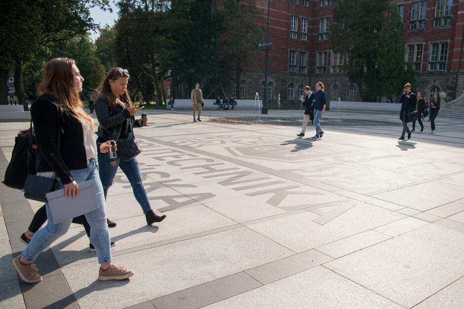 Ludzie chodzą po placu przed gmachem uczelni. Na placu duży napis Politechnika Gdańska .