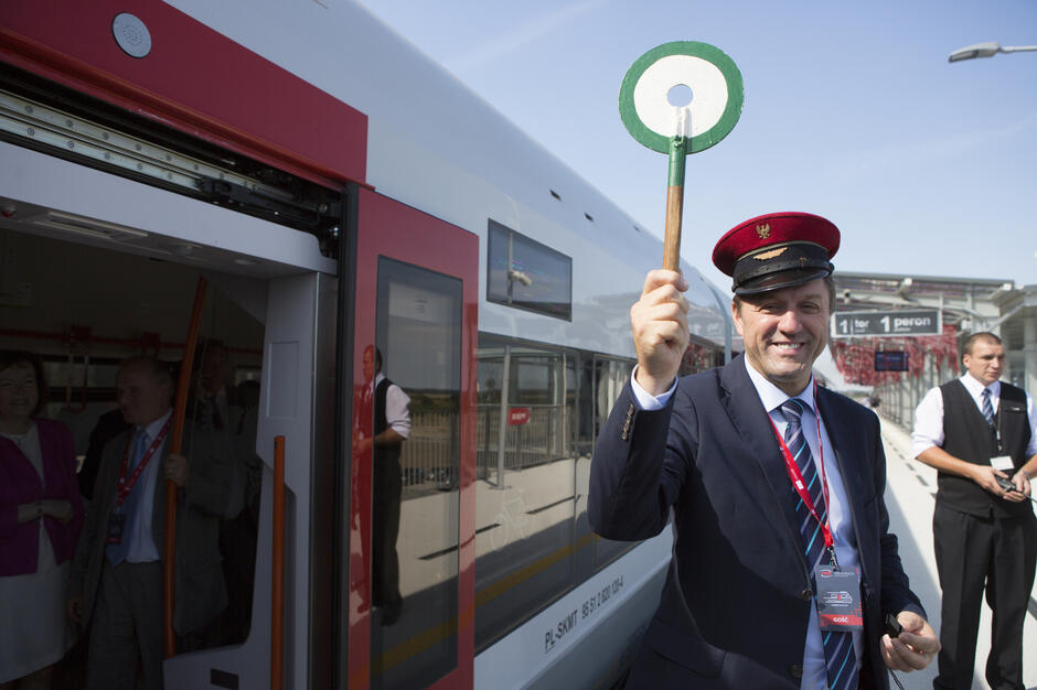Na zdjęciu widać uśmiechniętego mężczyznę w kolejarskim mundurze i czerwonej czapce, trzymającego w górze lizak do dawania sygnałów pociągom. W tle stoi nowoczesny skład kolejowy z otwartymi drzwiami oraz kilka osób na peronie. 