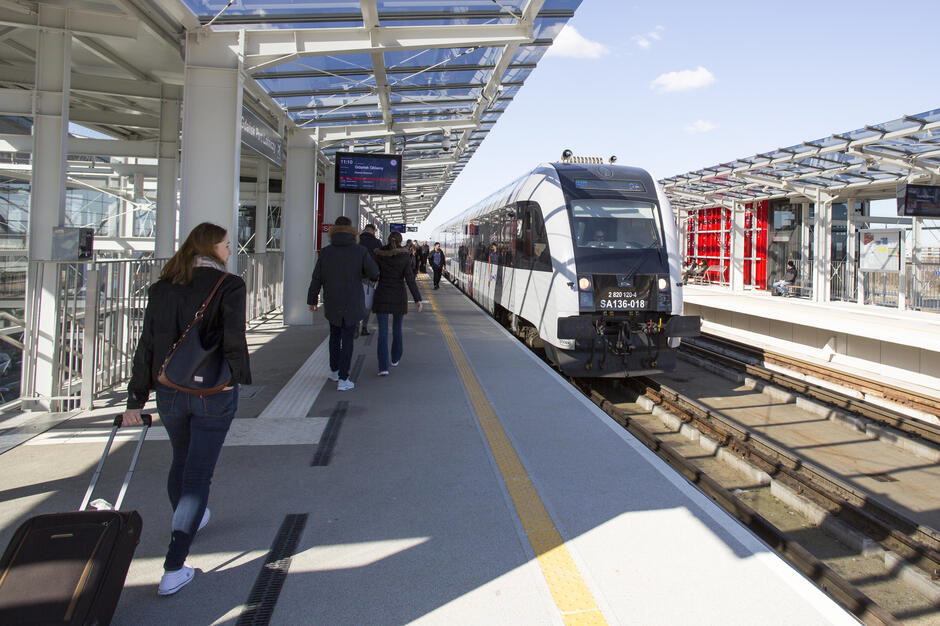 Na zdjęciu widać nowoczesny pociąg stojący na peronie stacji kolejowej, do którego podchodzą pasażerowie. Na pierwszym planie kobieta ciągnie walizkę na kółkach, zmierzając w kierunku pociągu. 