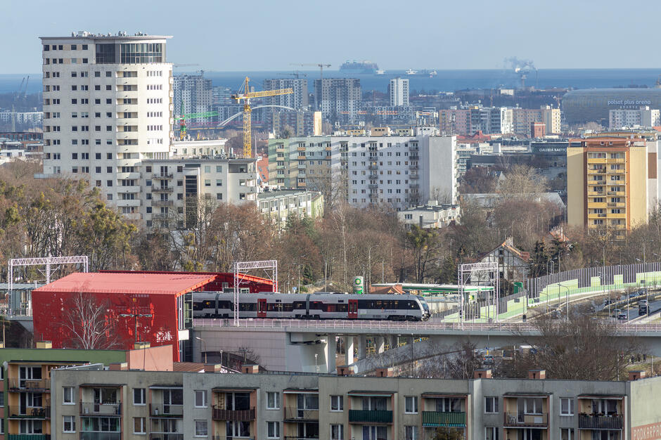 Na zdjęciu widać pociąg PKM przejeżdżający przez estakadę obok charakterystycznego czerwonego budynku stacji Niedźwiednik w Gdańsku. W tle rozciąga się panorama miasta z wysokimi blokami, żurawiami budowlanymi i widocznym w oddali Morzem Bałtyckim. 