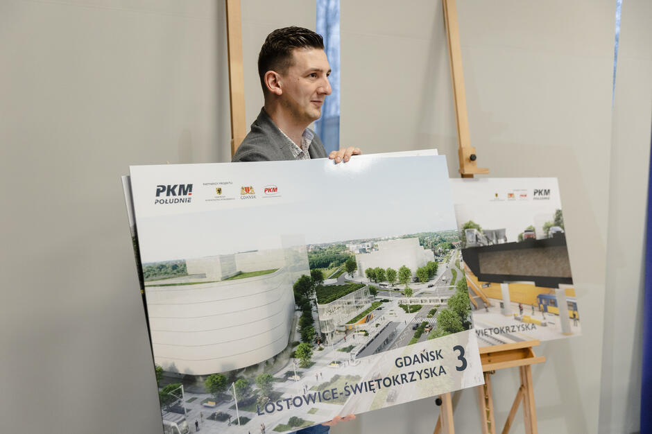 Na zdjęciu mężczyzna prezentuje planszę z wizualizacją projektu nowoczesnej infrastruktury w ramach inwestycji PKM Południe. Na planszy widnieje napis „Gdańsk Łostowice-Świętokrzyska” oraz logo partnerów projektu. 