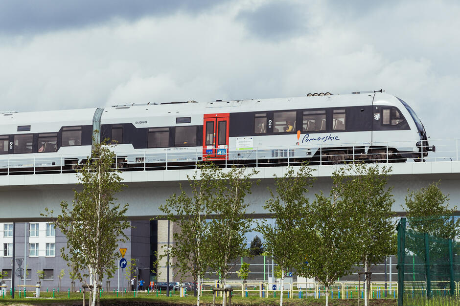 Na zdjęciu widać nowoczesny pociąg jadący po estakadzie, oznakowany logo „Pomorskie”. W tle znajdują się budynki mieszkalne i drzewa, a pod wiaduktem przebiega ulica z samochodami. 