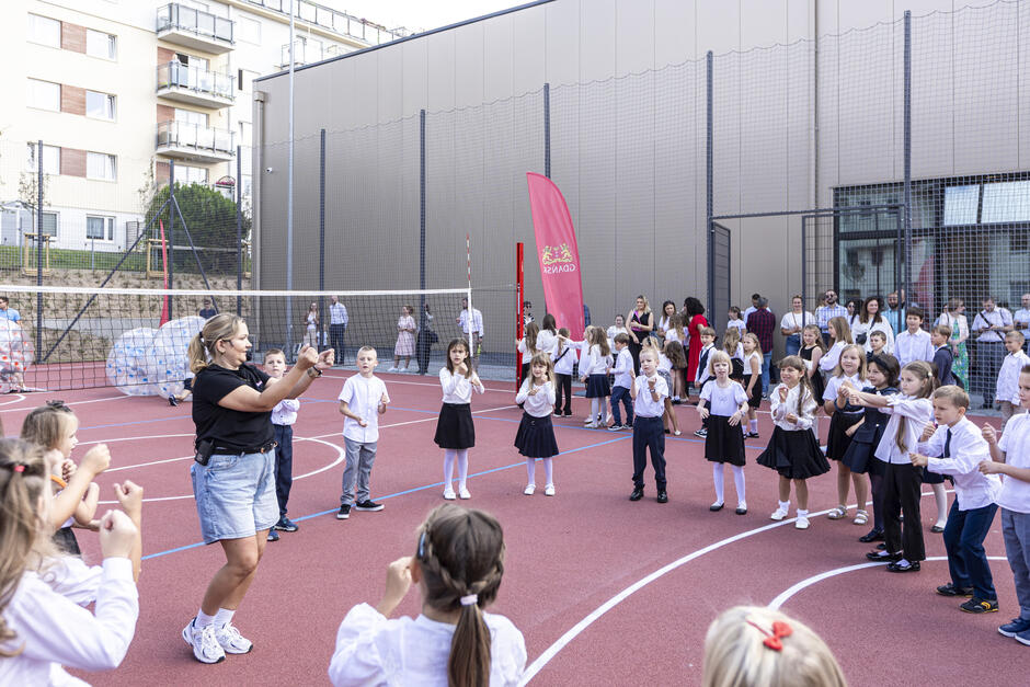 na boisku szkolnym stoi grupka uczniów w galowych strojach, stoją w okręgu, patrzą na nauczycielkę