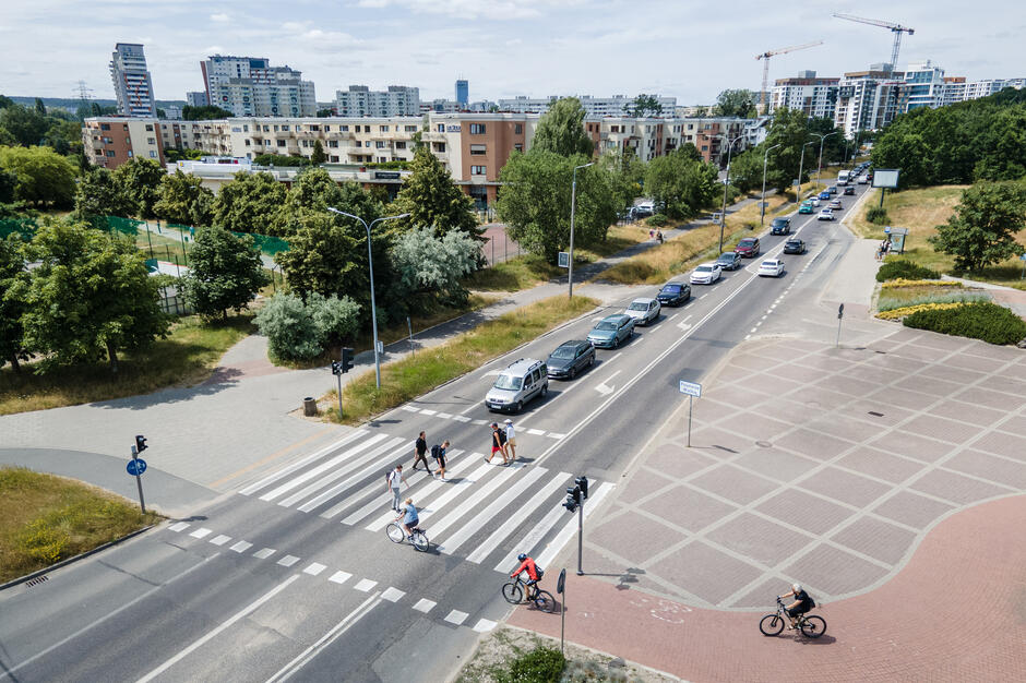 Na zdjęciu widać ruchliwą ulicę z przejściem dla pieszych, po którym przechodzą ludzie i przejeżdżają rowerzyści. W tle stoją samochody w korku, a za nimi widoczne są osiedla mieszkaniowe oraz żurawie budowlane.
