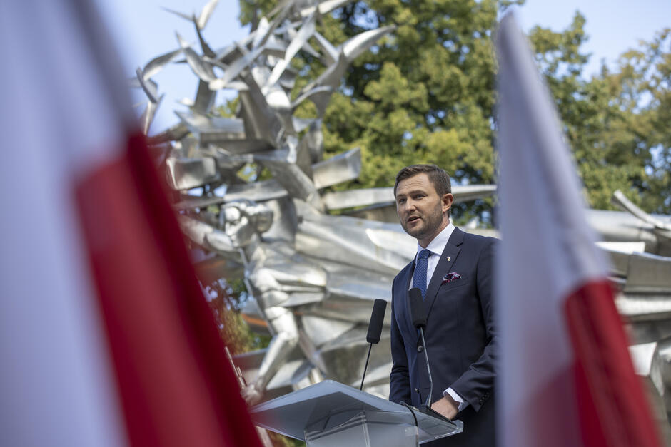 Mężczyzna w garniturze przemawia na mównicy pod pomnikiem, między flagami polskimi.