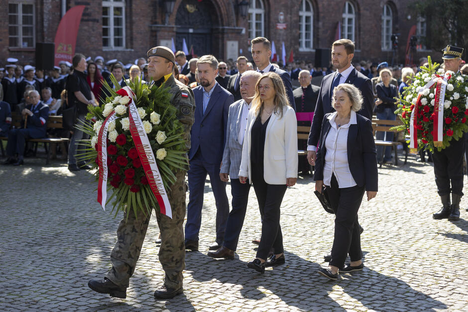 Kilka osób idzie za mężczyzną w mundurze niosącym wieniec by złożyć kwiaty pod pomnikiem.