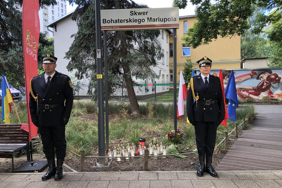 Znicze stoją na ziemi, pod szyldem z nazwą skweru. Po bokach na baczność stoi dwoje strażników - po lewej mężczyzna, po prawej - kobieta