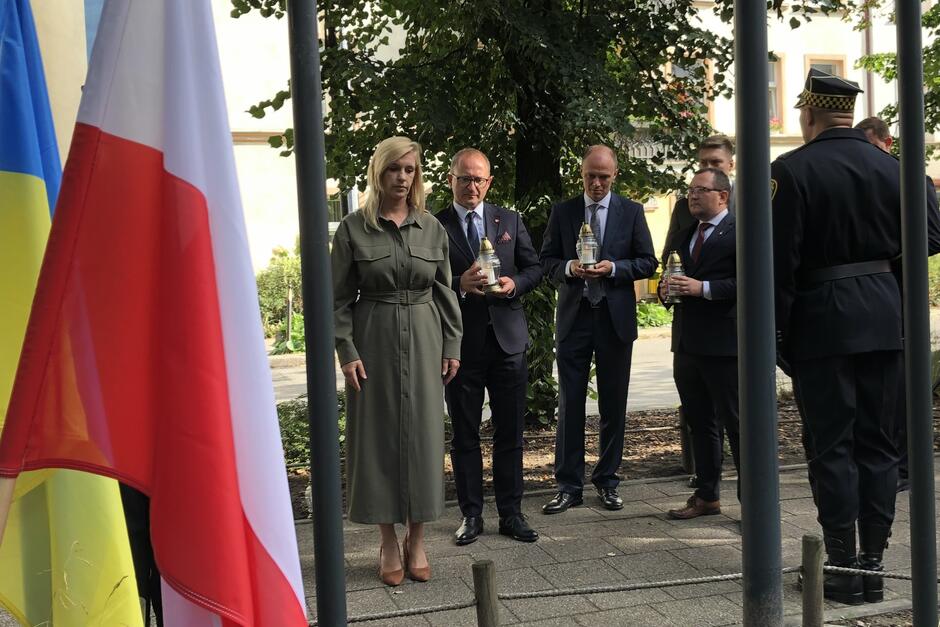 Cztery osoby w trakcie składania zniczy w uroczystym miejscu. Pierwsza od lewej jest kobieta, na prawo od niej są trzej mężczyzźni, każdy trzyma szklany znicz w rękach  