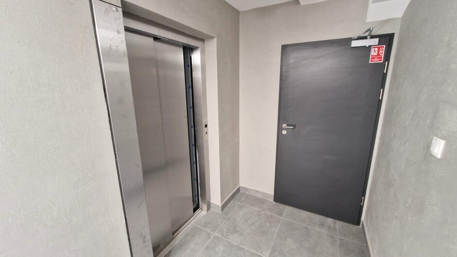 na zdjęciu korytarz z szarymi ścianami, po lewej widać drzwi windy, na wprost tradycyjne drzwi