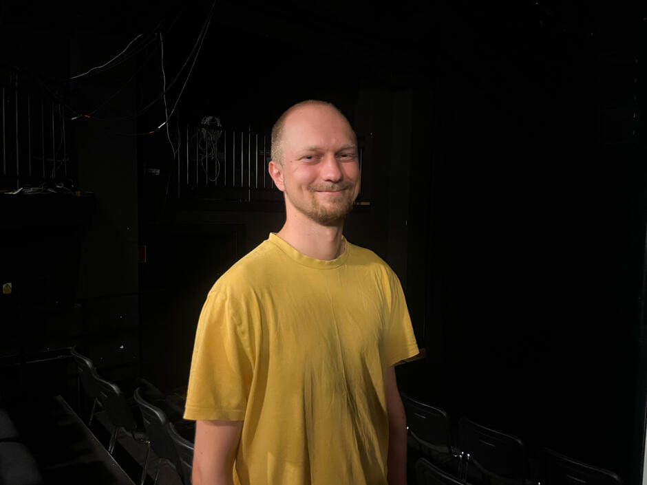 mężczyzna na czarnym tle, w żółtej koszulce, uśmiechnięty, z bardzo krótkimi włosami i lekkim zarostem