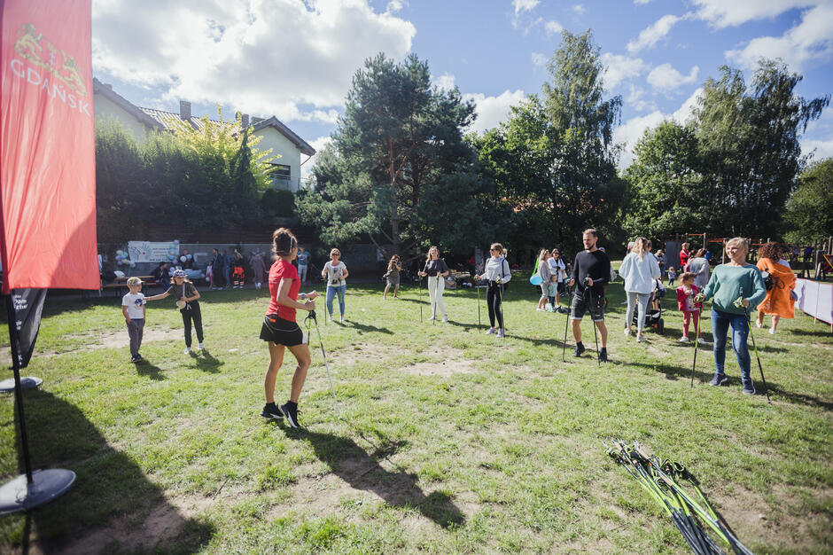 na zdjęciu młoda trenerka pokazuje ćwiczenie grupie stojącej wokół niej, na świeżym powietrzu, na trawie