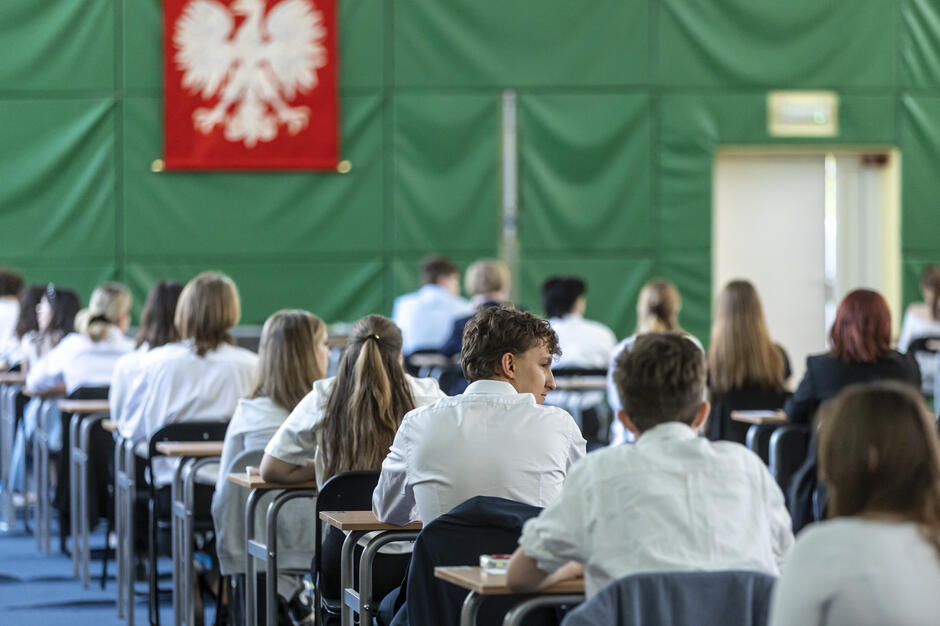 Na zdjęciu widać uczniów siedzących w sali egzaminacyjnej, ustawionych w równych rzędach przy oddzielnych ławkach. Na ścianie wisi godło Polski, co sugeruje, że to egzamin maturalny lub inny oficjalny test w polskiej szkole. 