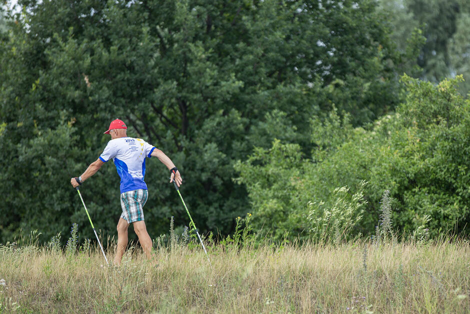 na zdjęciu mężczyzna idący z kijkami do nordic walking, jest w stroju sportowym idzie po zielonej łące, w tle zielony las