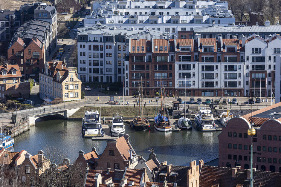 Zdjęcie ukazuje fragment Gdańska z nowoczesnymi apartamentowcami nad Motławą, przy których zacumowane są jachty i żaglówki. Na pierwszym planie widoczne są dachy zabytkowych kamienic, tworzące kontrast z nową zabudową.

