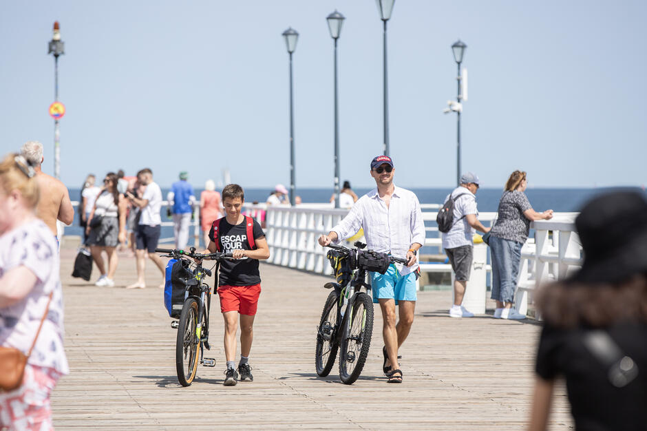na zdjęciu fragment drewnianego mola, po którym spacerują ludzi, na pierwszym planie dwóch rowerzystów prowadzi rowery