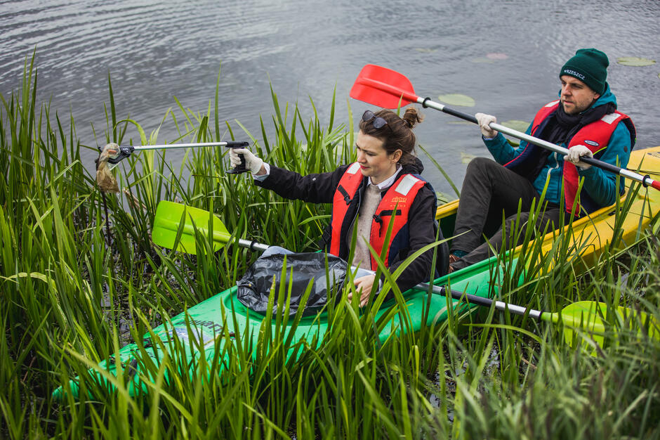 Kajak stoi w szuwarach. W kajaku z tyłu mężczyzna w czapce, z przodu kobieta ze szczypcami w ręku, którymi zbiera z rzeki śmieci. Między nogami kobieta ma worek na śmieci.