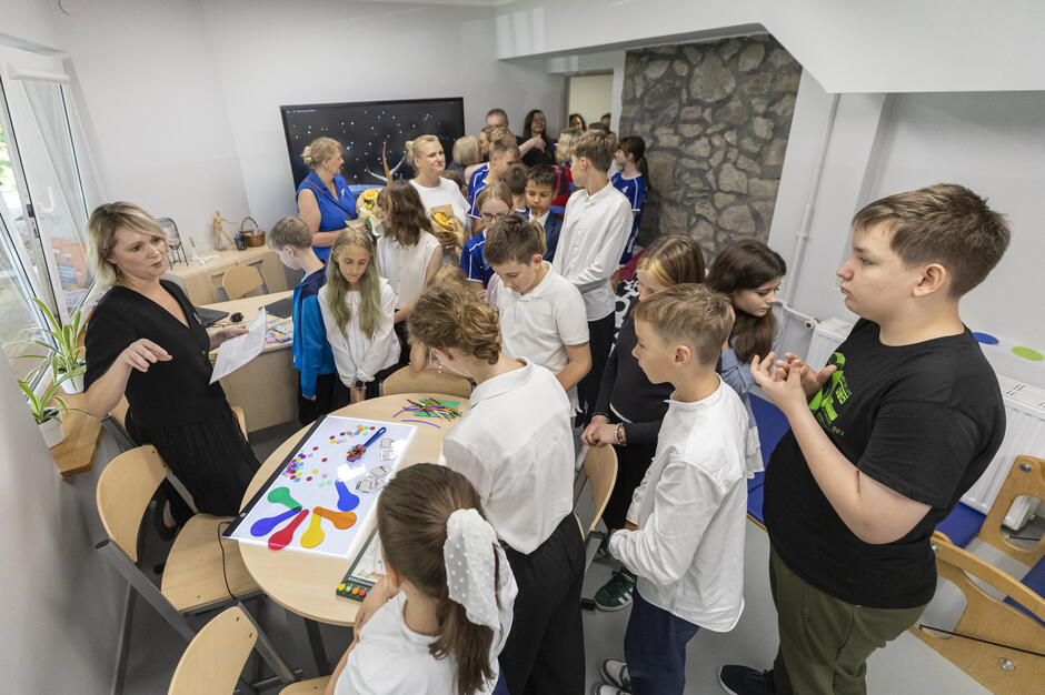 Na zdjęciu widać grupę uczniów zgromadzonych wokół stołu z kolorowymi materiałami edukacyjnymi, prowadzonych przez nauczycielkę. W tle stoją pozostali uczniowie oraz dorośli, obserwując przebieg zajęć w nowoczesnej sali. 
